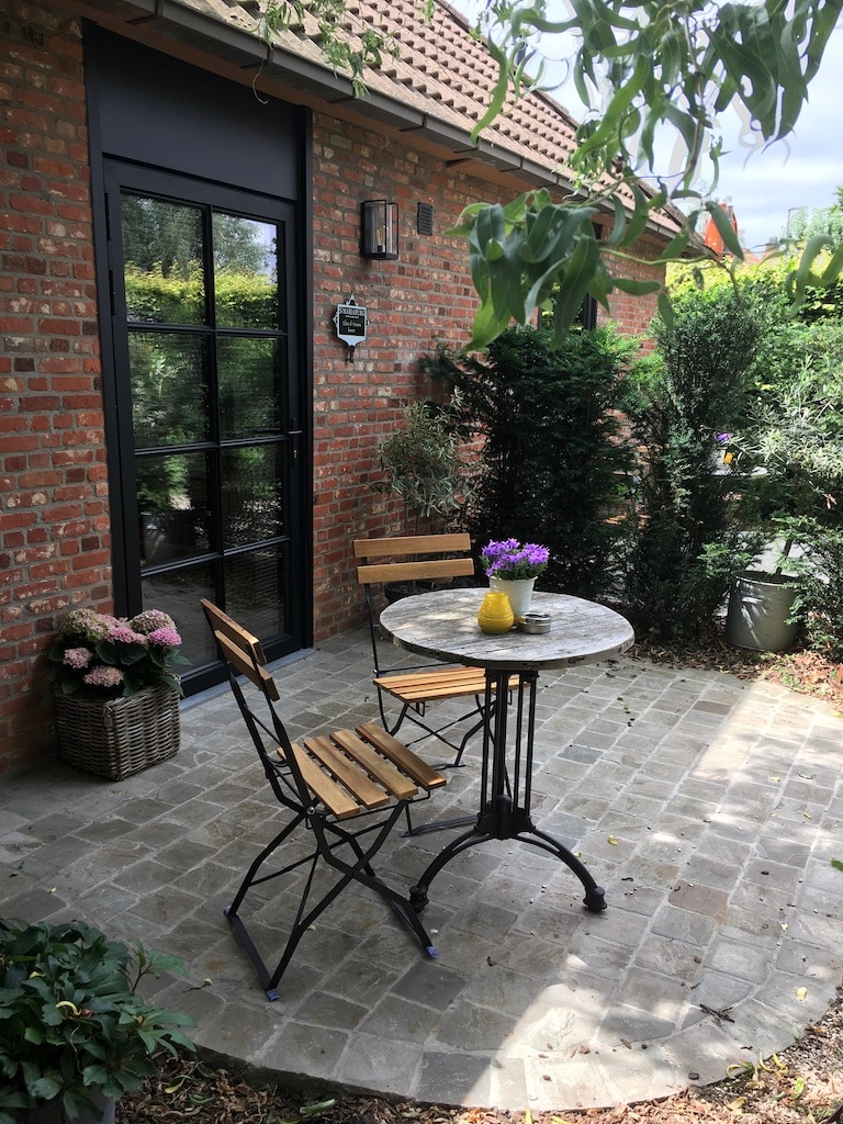 A private terrace features a round stone table surrounded by two wooden chairs. Potted plants and flowers add greenery to the setting, while a brick wall provides a rustic backdrop. The entrance includes large glass doors, allowing natural light into the area.