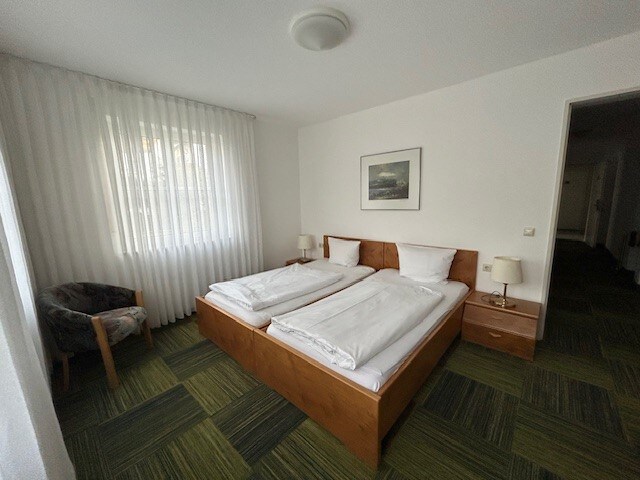 A well-lit bedroom features two spacious beds arranged side by side, each fitted with white bedding. A framed artwork is displayed on the wall above the bed. Natural light filters through sheer white curtains. A cozy armchair and bedside tables with lamps complement the room.