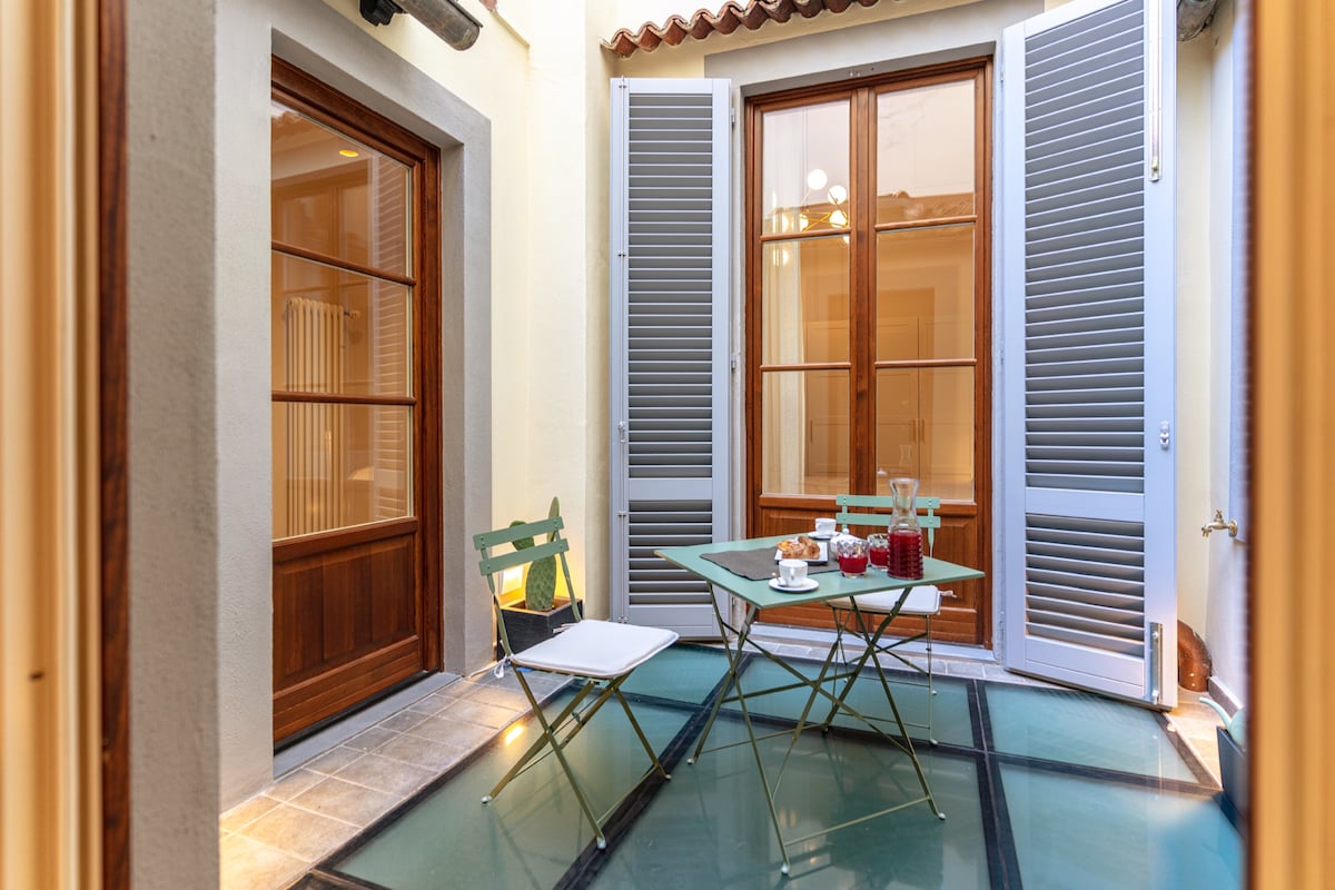 A charming outdoor space is presented with light-colored walls and two large wooden windows flanked by grey shutters. A small glass table with two folding chairs is set with refreshments, inviting relaxation in the private courtyard.