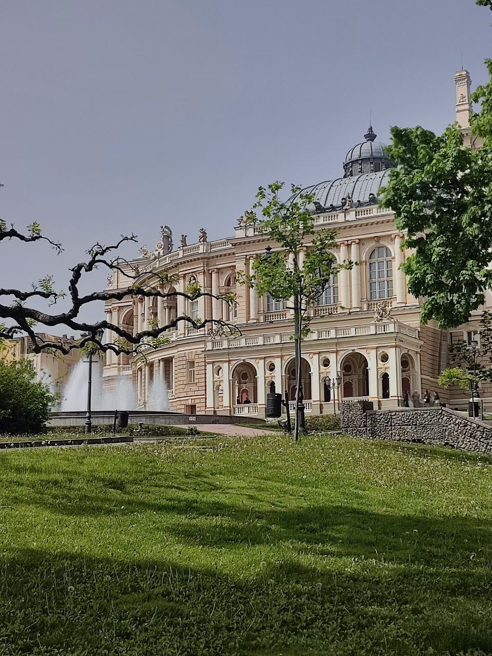 The image showcases an elegant building with a grand façade and a domed roof, surrounded by lush greenery. A fountain can be seen in front of the structure, adding a serene water element to the scene. Tall trees provide shade in this tranquil environment.