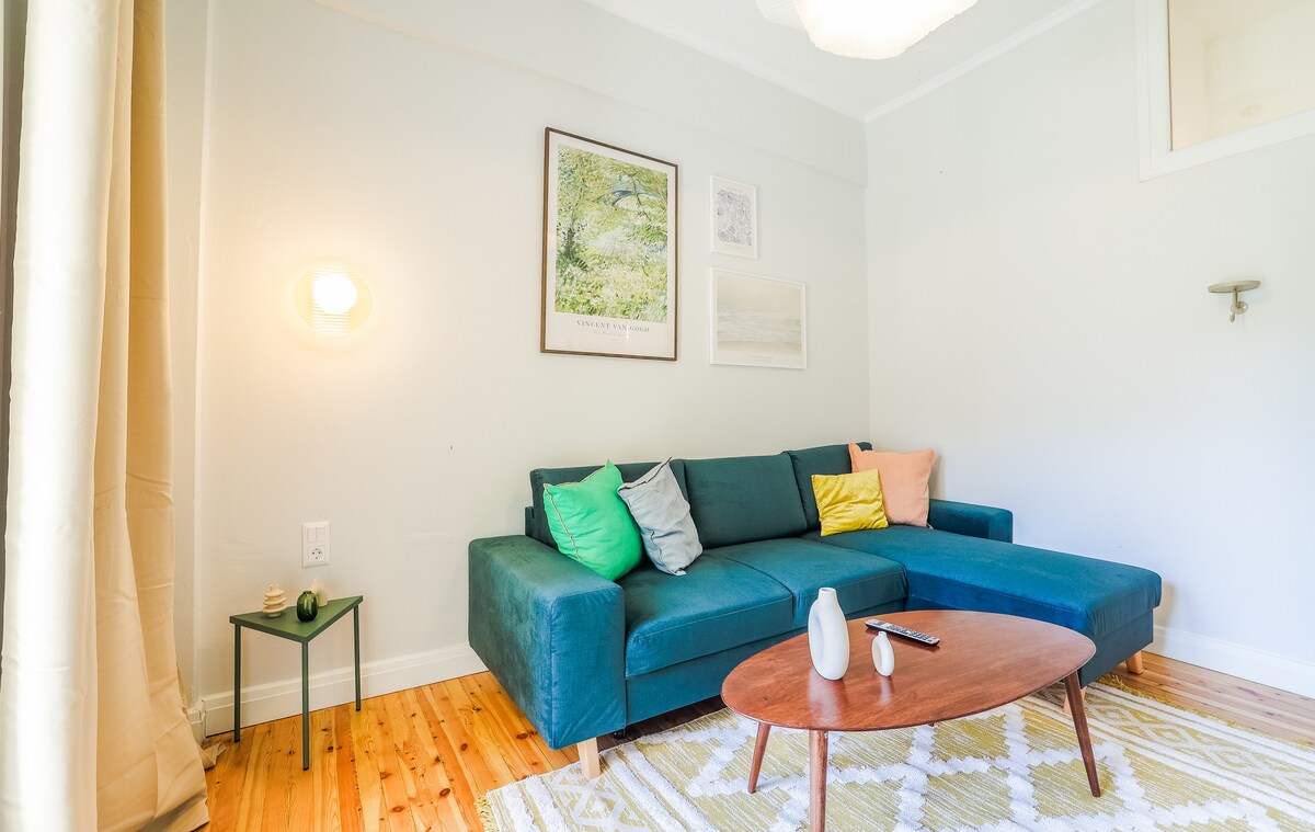 A modern living area features a spacious green sectional sofa adorned with decorative pillows in various colors. A round wooden coffee table is positioned in front, and a small side table holds a minimalist vase. Light walls create an airy ambiance, complemented by soft lighting.