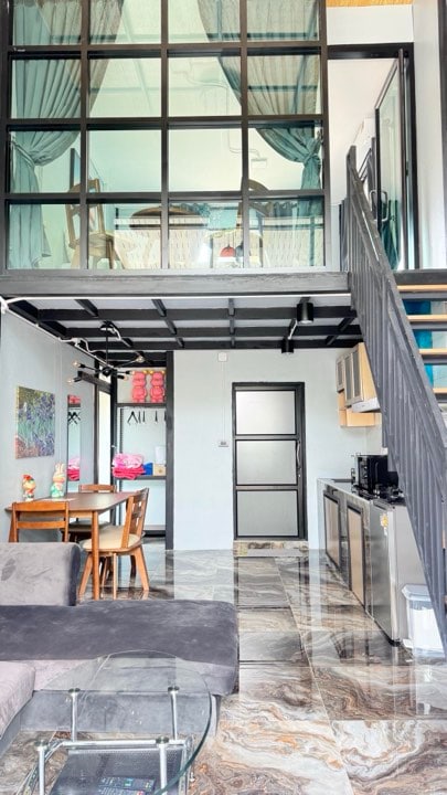 A modern living area showcases a gray sectional sofa beside a stylish glass coffee table. A dining table with wooden chairs is positioned across from an open kitchen equipped with stainless steel appliances. A staircase leads to an upper level, with dark-framed glass providing a view of the space above.
