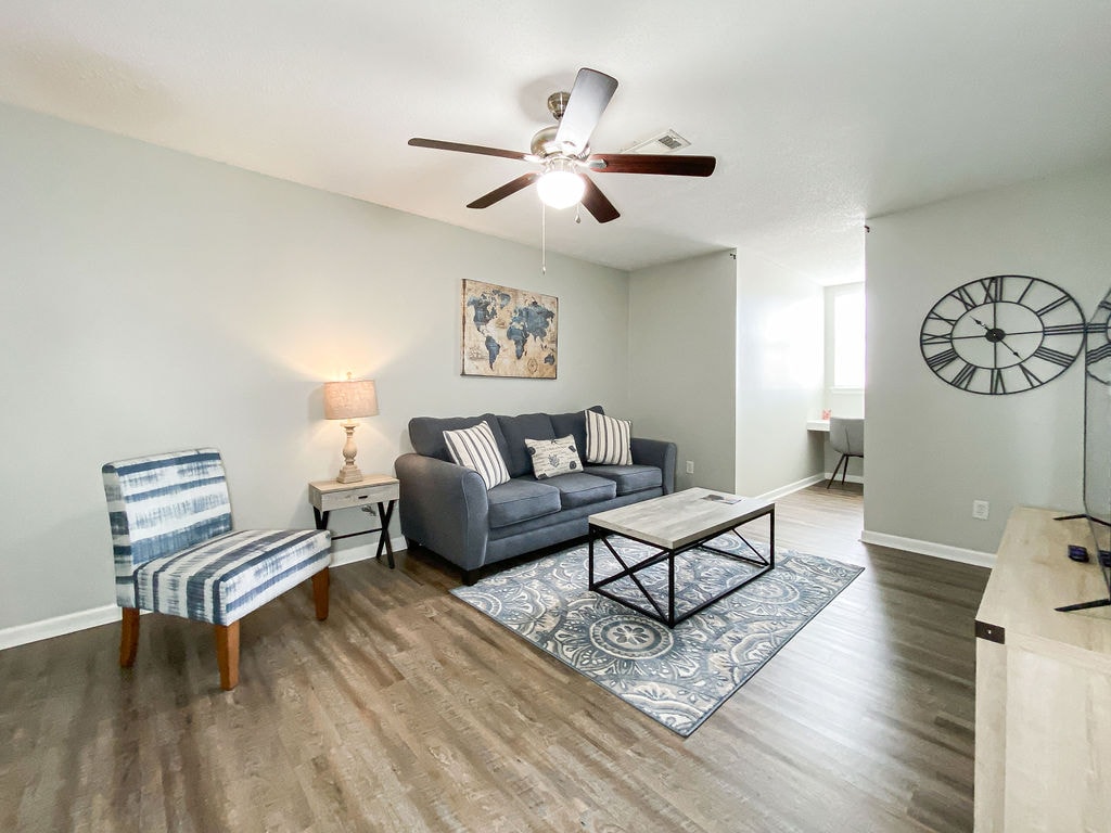 A comfortable living room area features a gray sofa with accent pillows, a striped chair, and a coffee table on a decorative rug. A wall-mounted clock and a piece of wall art add to the decor. Natural light streams through a nearby window.