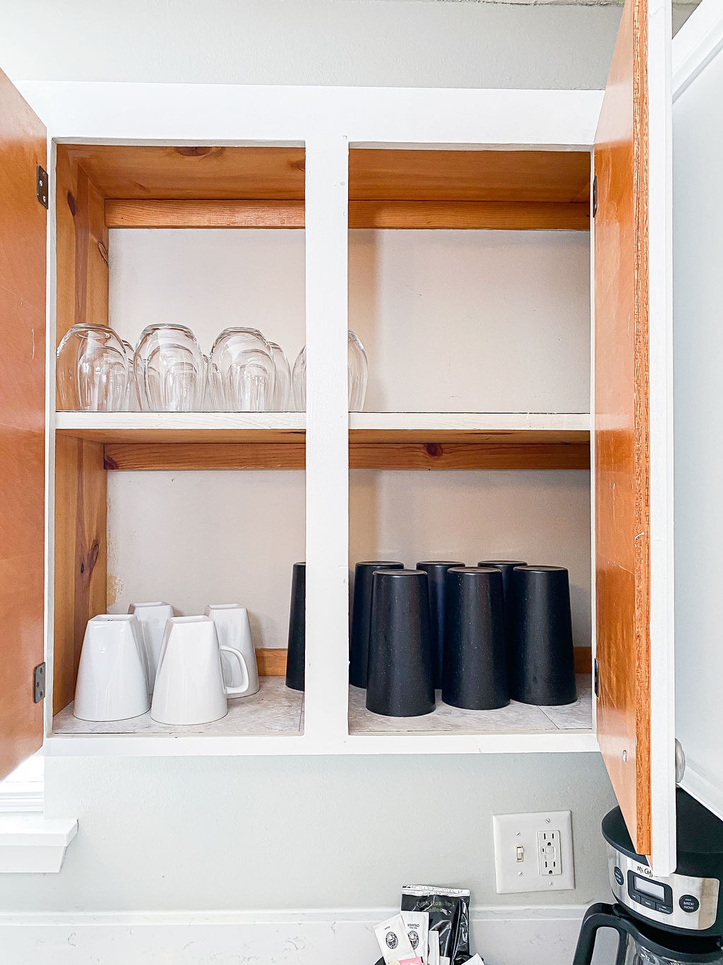 A kitchen cabinet is shown with two shelves displaying a set of clear glasses on the top and a collection of white and black mugs below. The items are neatly arranged, contributing to an organized kitchen environment.