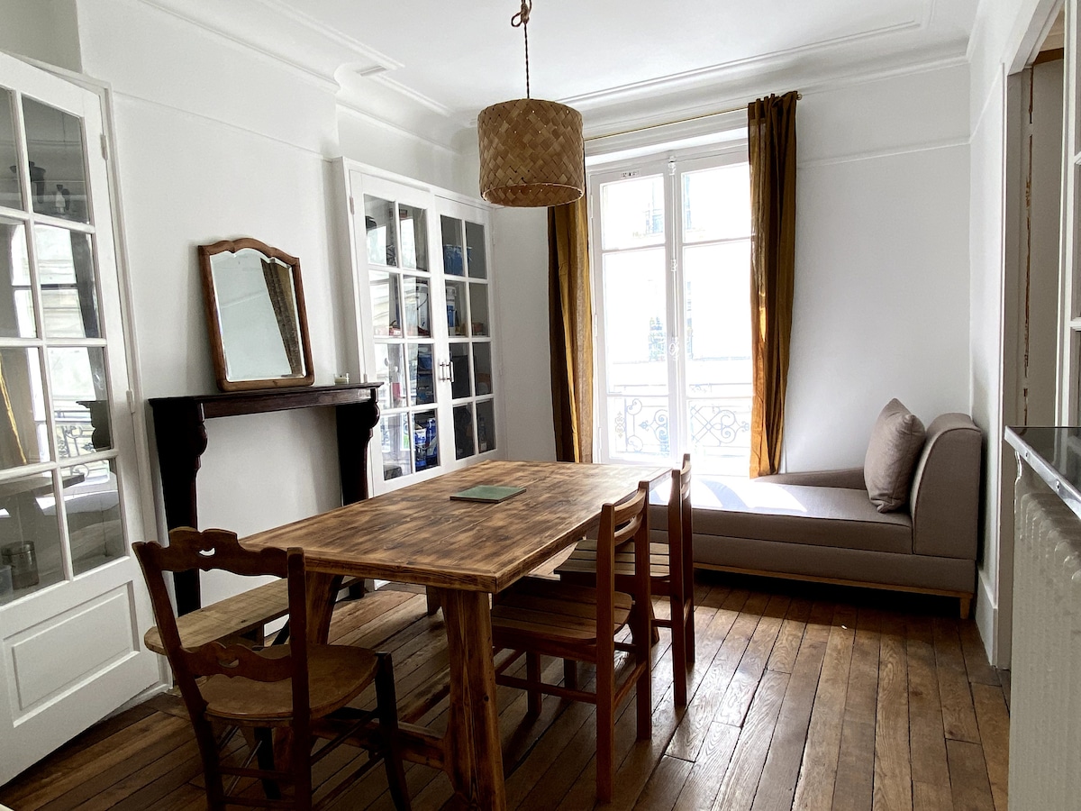 A dining area features a wooden table surrounded by four chairs and an inviting window seat. Natural light enters through large windows adorned with elegant curtains. A mirror is placed above a traditional fireplace, complementing the hardwood flooring throughout the space.