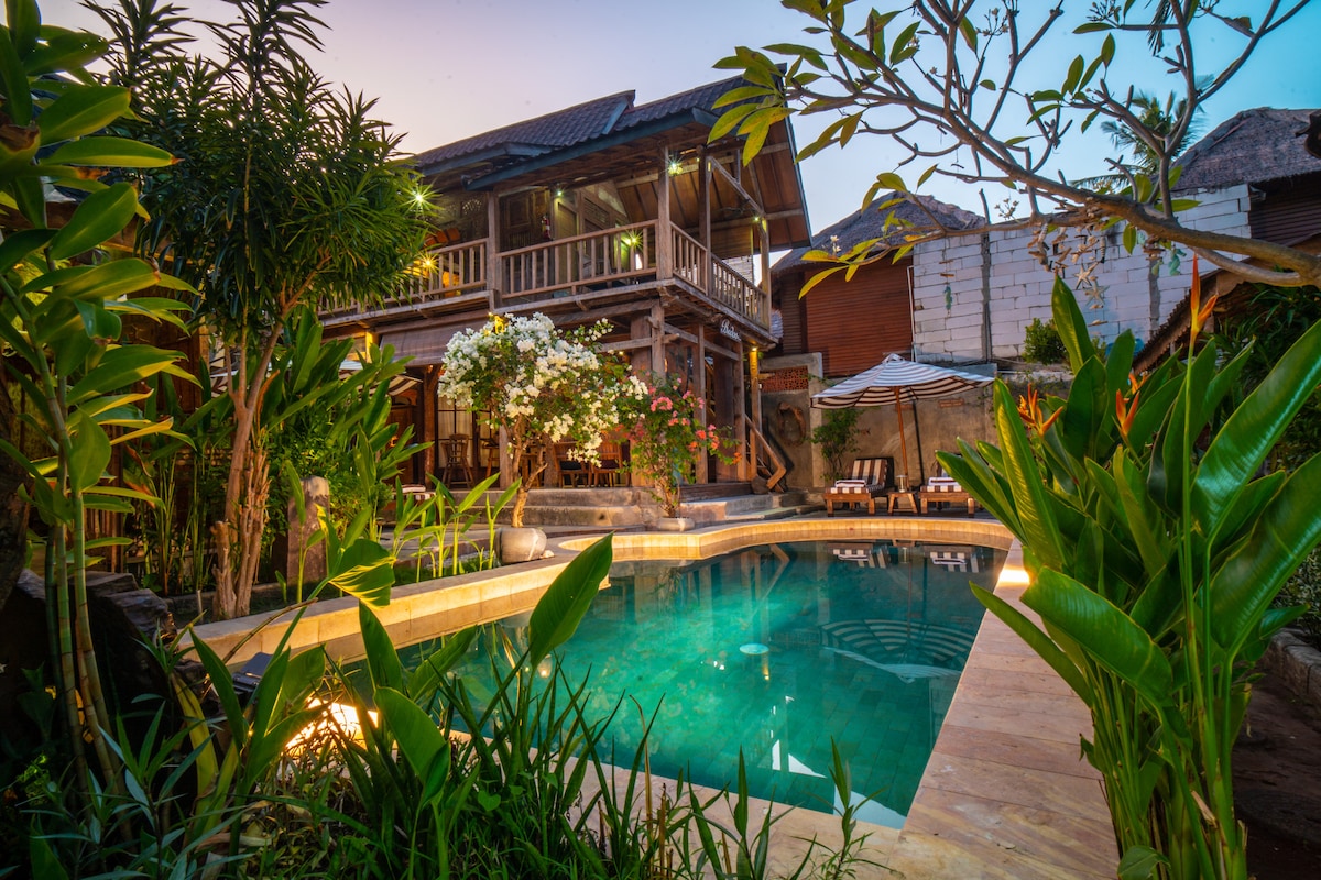 A private villa is surrounded by lush greenery, featuring a serene swimming pool framed by tropical plants. The wooden structure showcases a charming design with multiple levels, while soft lighting accentuates the inviting atmosphere during twilight.