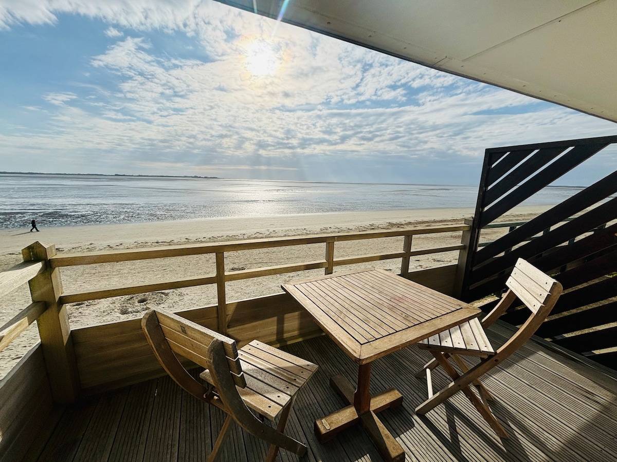 A wooden balcony features a table and two chairs, providing a serene space to enjoy views of the beach and sea. The horizon showcases shimmering water reflecting sunlight, while fluffy clouds drift across the sky.