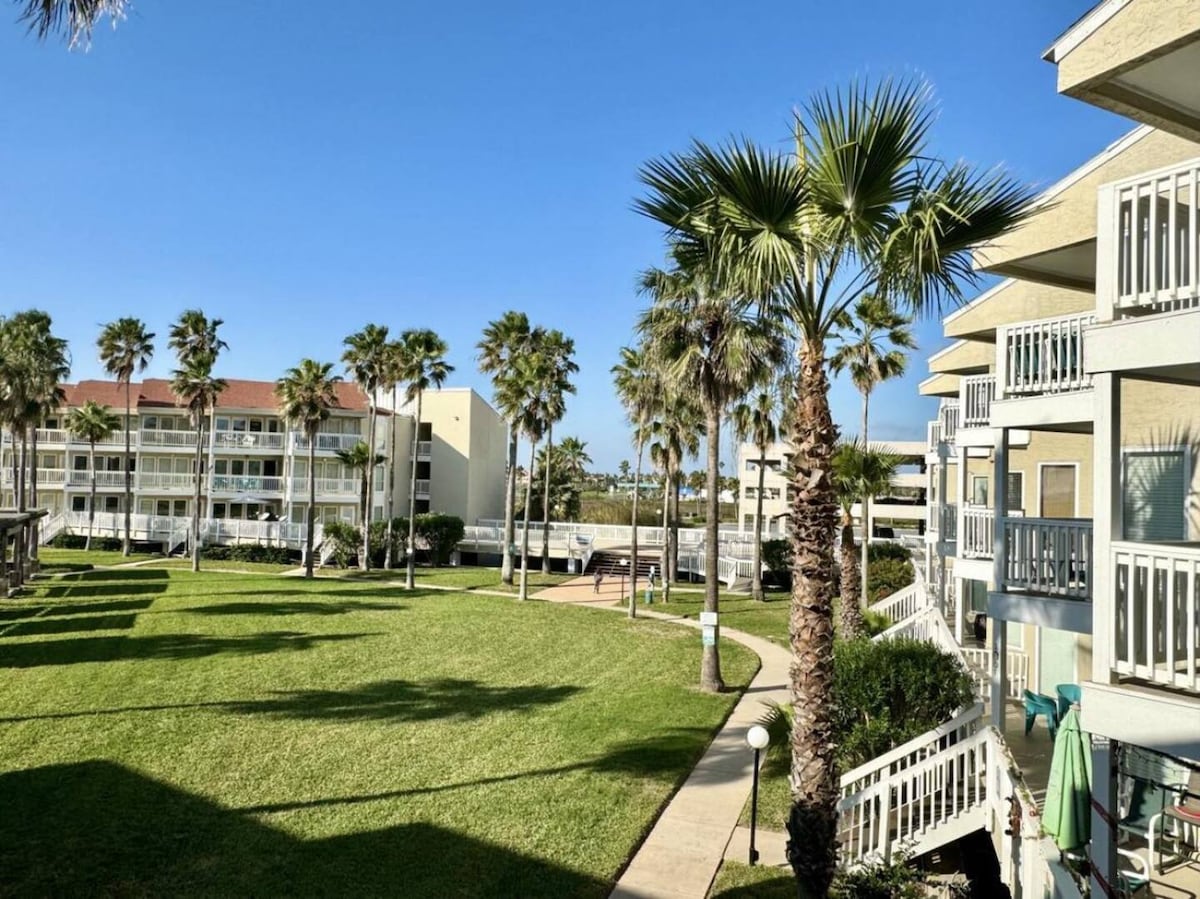 A landscaped courtyard is visible, featuring well-maintained grass and several palm trees. Surrounding structures include multiple apartment buildings, each with balconies. The clear blue sky adds to the serene environment, making the outdoor space inviting and open.