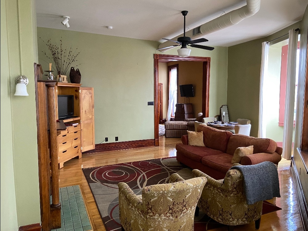 A spacious living room features a combination of warm green walls and hardwood floors. A large area rug with intricate patterns anchors the seating arrangement, which includes a comfortable sectional and two patterned chairs. Natural light streams through the large windows, enhancing the inviting atmosphere.