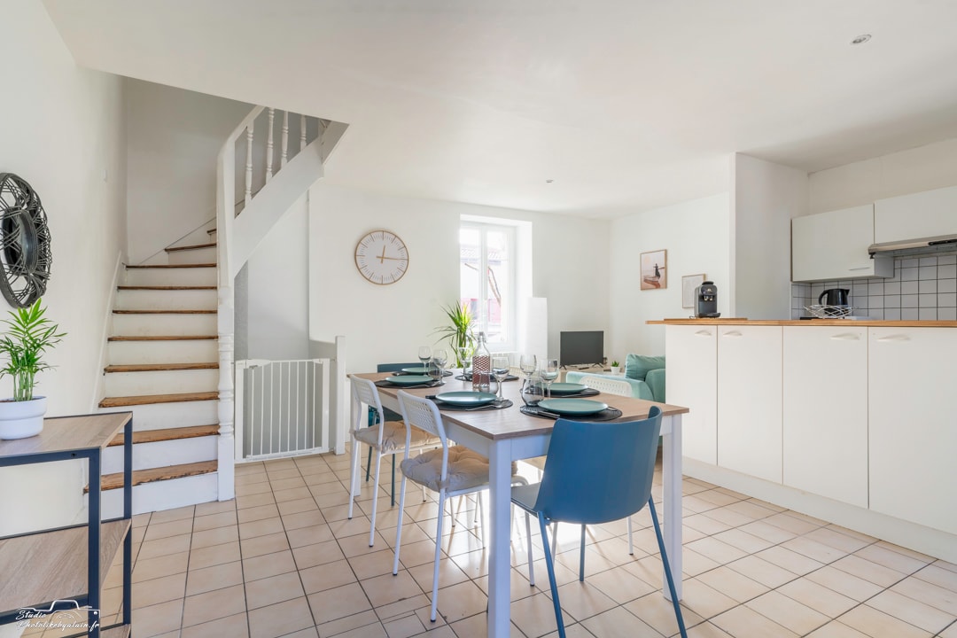 A bright open-plan kitchen and living area features a dining table set for four, with modern chairs and a decorative plant. A staircase leads to the upper floor, while a wall clock and television enhance the space's functionality.