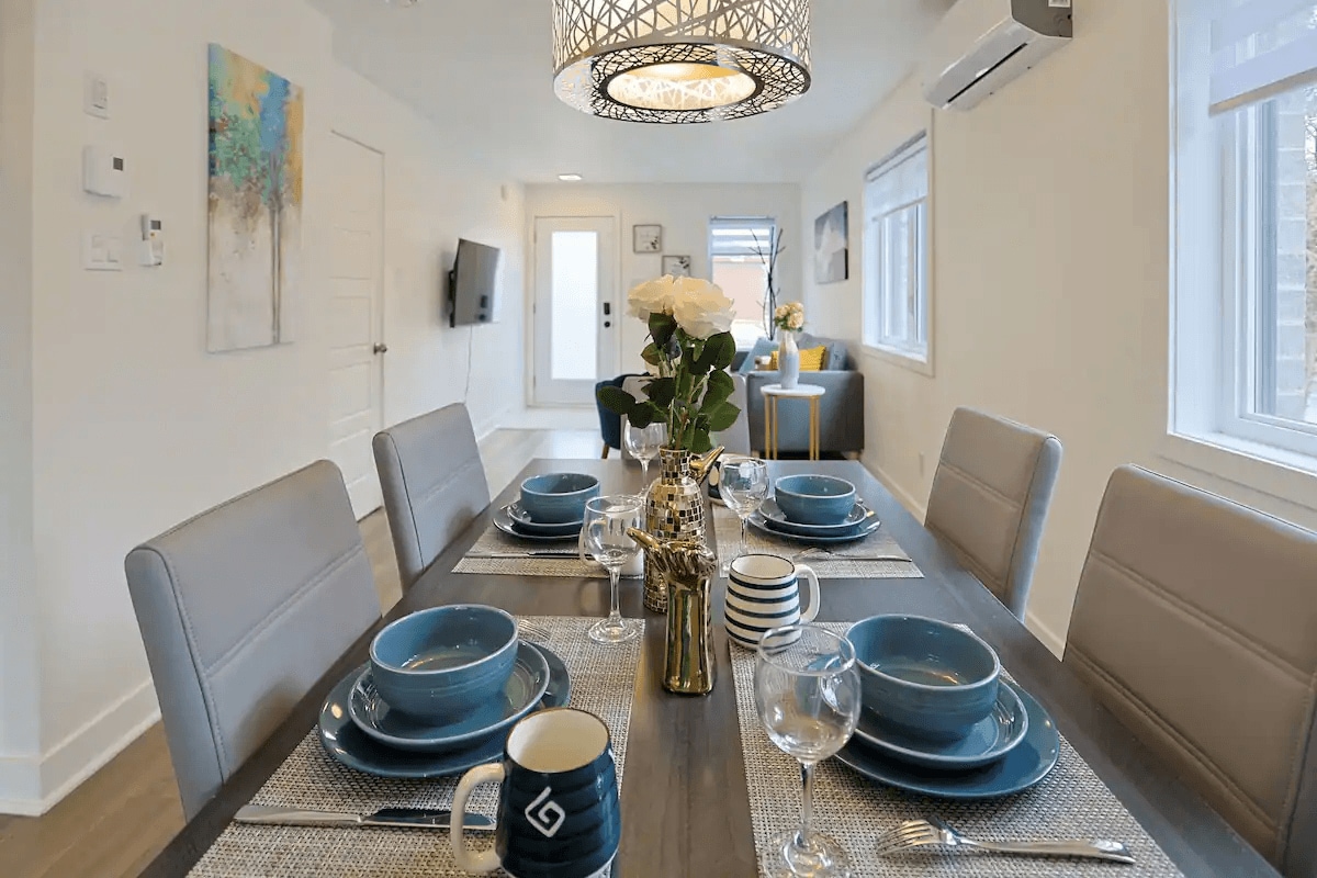 A modern dining area is presented, featuring a spacious table set with stylish dishware. Blue bowls are arranged alongside sparkling glassware and a decorative vase with fresh flowers at the center. The room benefits from ample natural light, enhancing its inviting atmosphere.