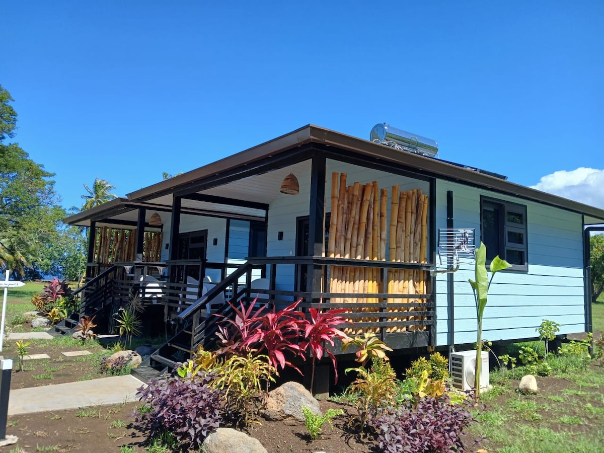 A single-story structure with a welcoming design is surrounded by lush greenery and tropical plants. The exterior features a combination of light blue and natural wood accents, with a covered porch supported by textured columns. A clear sky and distant water view complement the setting.
