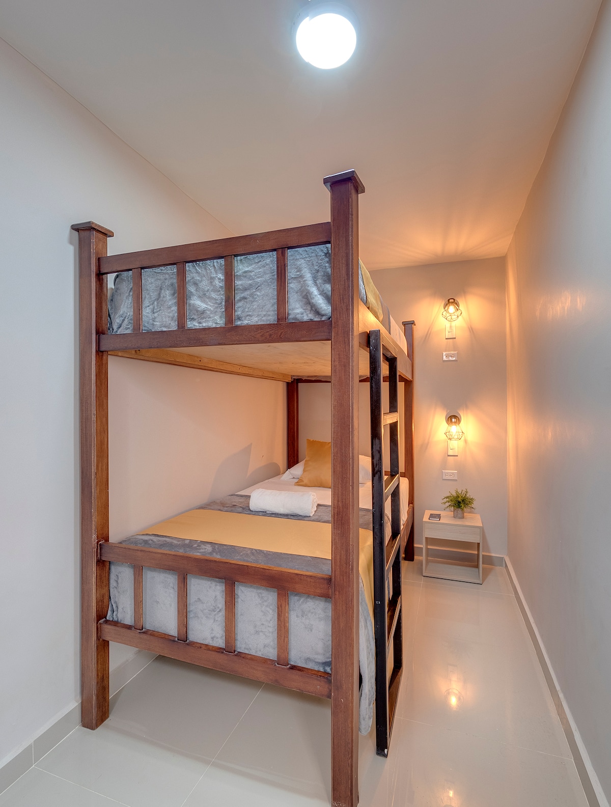A wooden bunk bed is positioned against a light-colored wall, featuring soft bedding in neutral tones. Warm pendant lights are mounted on the wall, illuminating the space. A small nightstand with a decorative plant is situated beside the lower bunk.