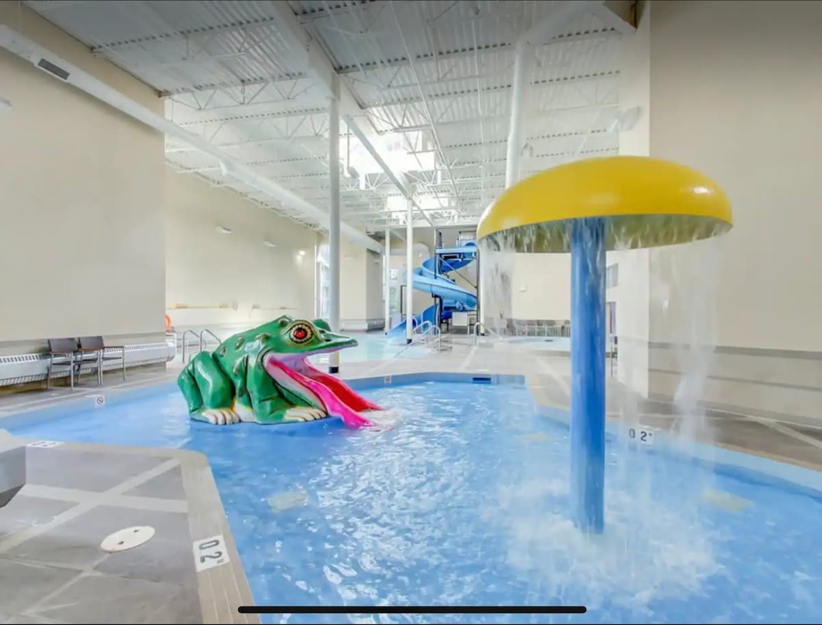 An indoor pool area features a colorful frog sculpture and a playful water feature resembling a mushroom, providing an engaging attraction for guests. A blue slide and spacious lounging areas are visible, ensuring accessibility and enjoyment for all ages.