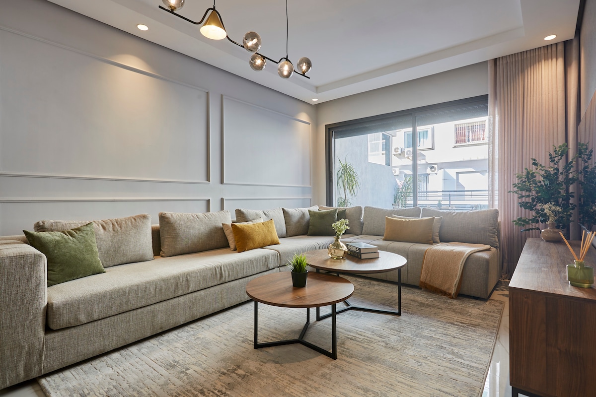 The spacious living area features a large, L-shaped sectional sofa adorned with decorative pillows. A coffee table sits at the center, accompanied by a small plant and a few decorative items. Natural light filters through the sliding glass doors, revealing a view of the outdoors.