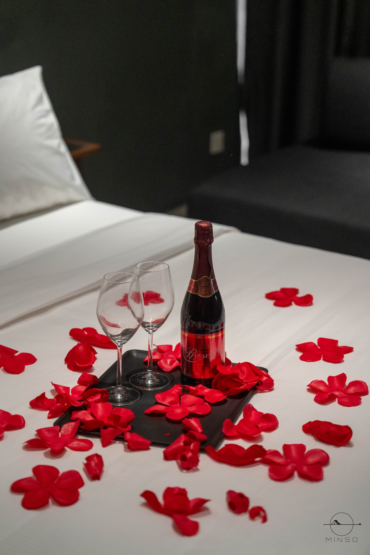 A set of two glasses and a bottle of sparkling beverage are arranged on a dark cloth, surrounded by scattered red rose petals on a smooth white bedspread. The bed is neatly made with white linens complemented by a decorative dark backdrop.