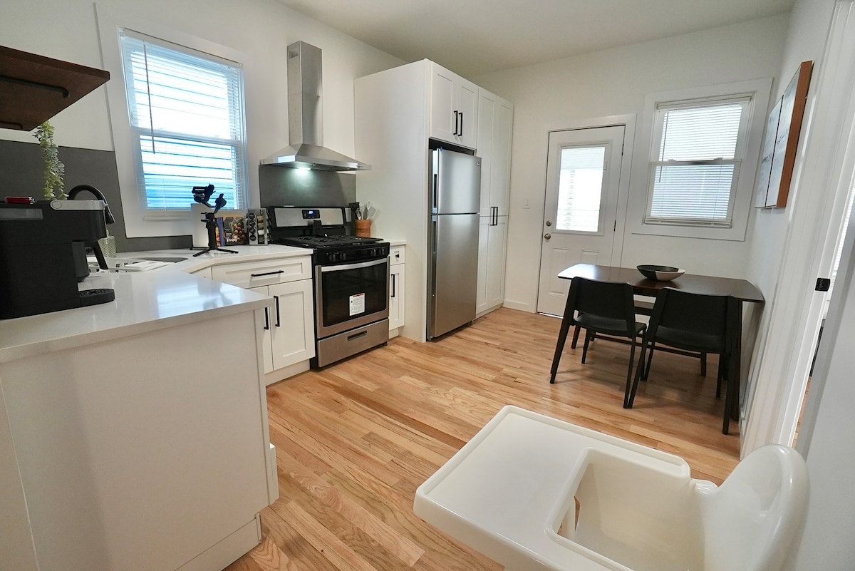 A modern kitchen space is shown with wooden flooring and sleek white cabinetry. Stainless steel appliances are present, including a refrigerator and stove. A dining area with a dark table and chairs is visible, alongside a high chair, creating a family-friendly atmosphere.