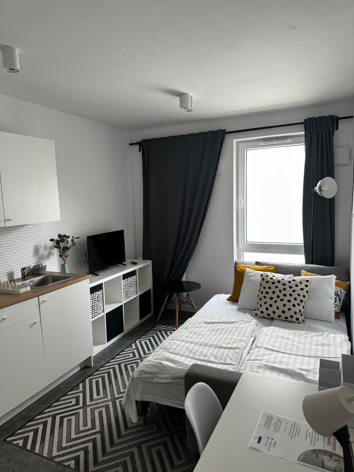 A compact living space features a cozy bed adorned with patterned cushions. A small kitchenette is visible with essential appliances. A mounted television is positioned near a black side table. Large windows are framed by dark curtains, allowing natural light to softly illuminate the room.