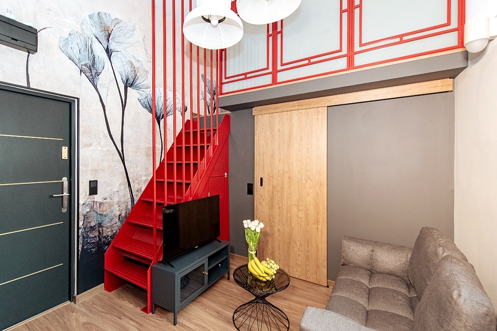 A compact living area features a gray sofa facing a television setup. A round table holds fresh fruits, and a floral wallpaper adds a touch of nature. A striking red staircase leads to a lofted sleeping area, enhancing the verticality of the space.