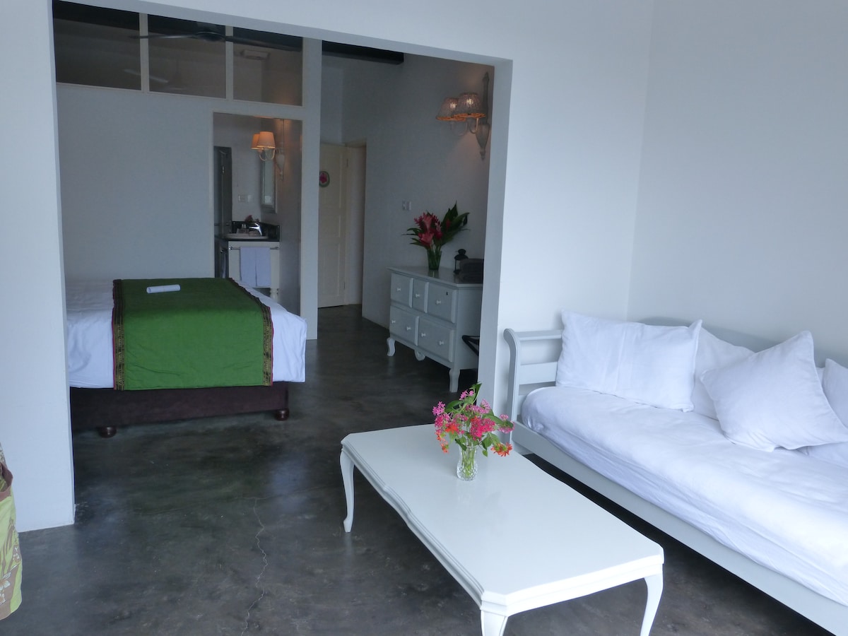 The interior space features a bright and open layout, with a spacious bed dressed in green linens and a complementary light sofa. A white coffee table adorned with fresh flowers sits in the center, enhancing the room's relaxed atmosphere. Soft lighting provides warmth throughout the area.