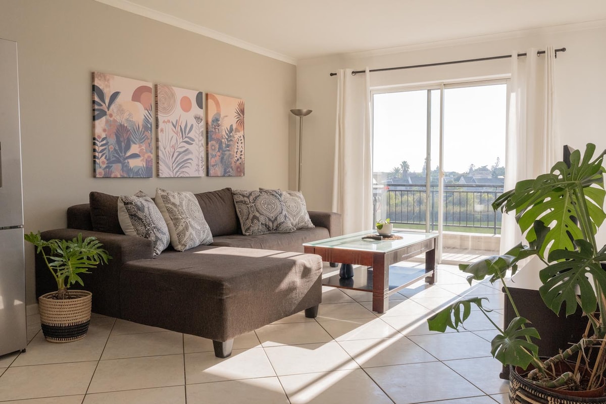 A bright lounge area features a large sectional couch adorned with decorative pillows. A glass coffee table sits in front of the couch, complemented by a potted plant. Natural light floods in through sliding glass doors, which open to a balcony overlooking the lake.