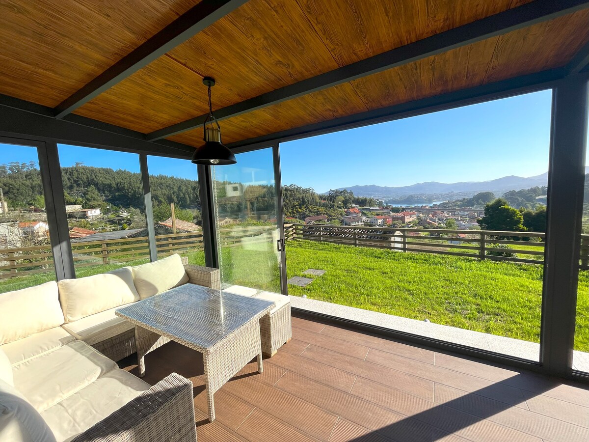 A sunlit porch features a comfortable seating area with a wicker sofa and a glass-top table. Large glass panels provide unobstructed views of the surrounding landscape, revealing lush greenery and distant hills. Natural light enhances the spacious feel of this inviting area.