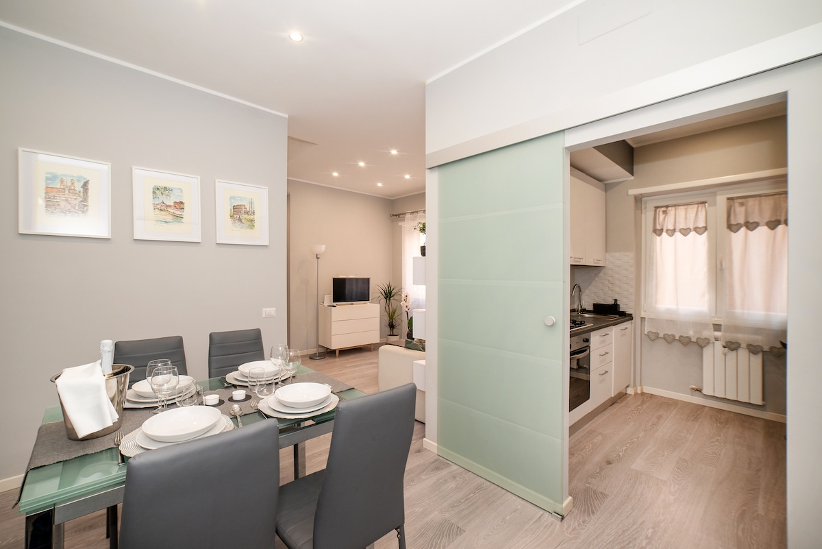 A dining area is visible, featuring a glass table set for five, with elegant tableware arranged neatly. Soft grey walls complement the space, while three framed artworks hang above the table. A sliding door leads to a neatly organized kitchen with natural light filtering through sheer curtains.