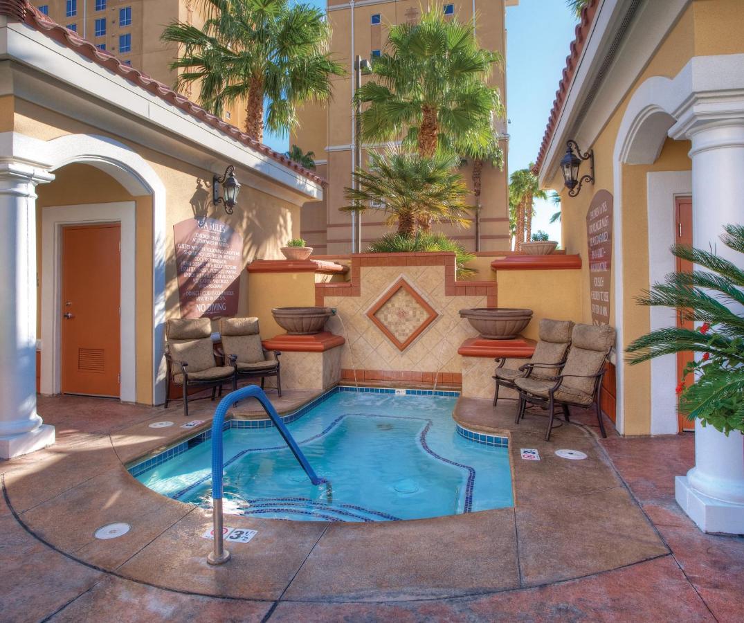 An outdoor hot tub is framed by tropical palm trees, surrounded by a tasteful patio area with two cushioned chairs. Decorative stone planters add visual interest, while a warm color palette complements the relaxed ambiance of this inviting space.