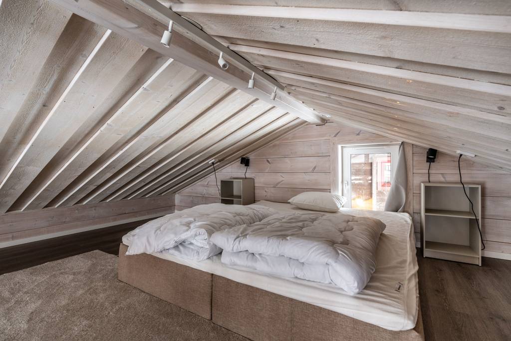 A loft area features a spacious double bed with layered bedding. The sloped wooden ceiling adds a cozy feel, while a window allows natural light to fill the room. Two bedside tables are positioned on either side of the bed, enhancing functionality.