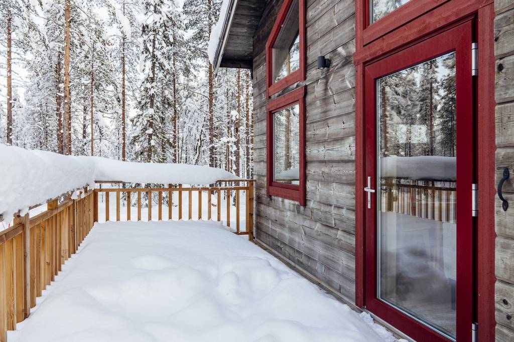 A wooden terrace is framed by a fence, surrounded by snow-covered ground and frosted trees in the background. Large windows with red frames provide a view into the cozy interior, enhancing the connection with the peaceful natural surroundings.