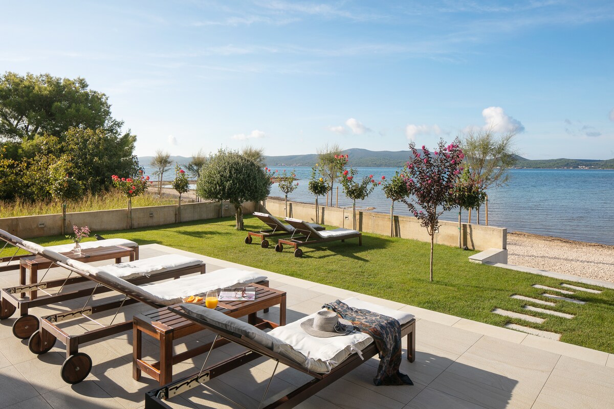 A landscaped garden space features sun loungers arranged on a patio, overlooking the sea. Vibrant greenery and flowering plants add color to the area. A stone pathway leads to the water’s edge, providing a tranquil view of the landscape and gentle waves.