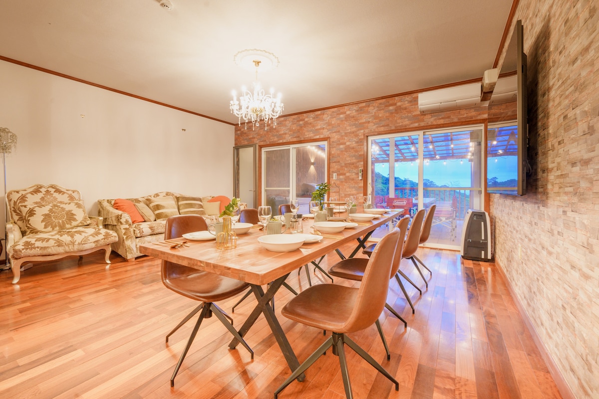 A dining area features a large wooden table set for dining, accompanied by several stylish chairs. A comfortable seating area with decorative sofas is visible in the background. Large glass doors provide a view of the outdoor patio, enhancing natural light in the space.