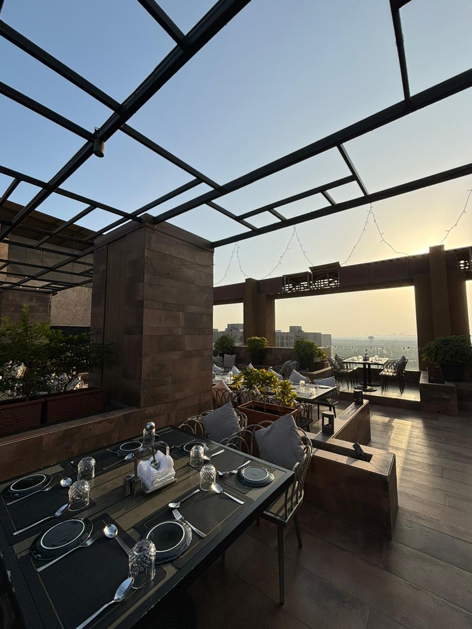 An outdoor dining space is presented under a metal-framed roof, featuring a polished table set for a meal. Surrounding greenery and seating create a relaxed ambiance. The horizon is visible in the background, with soft sunlight casting a warm glow over the scene.