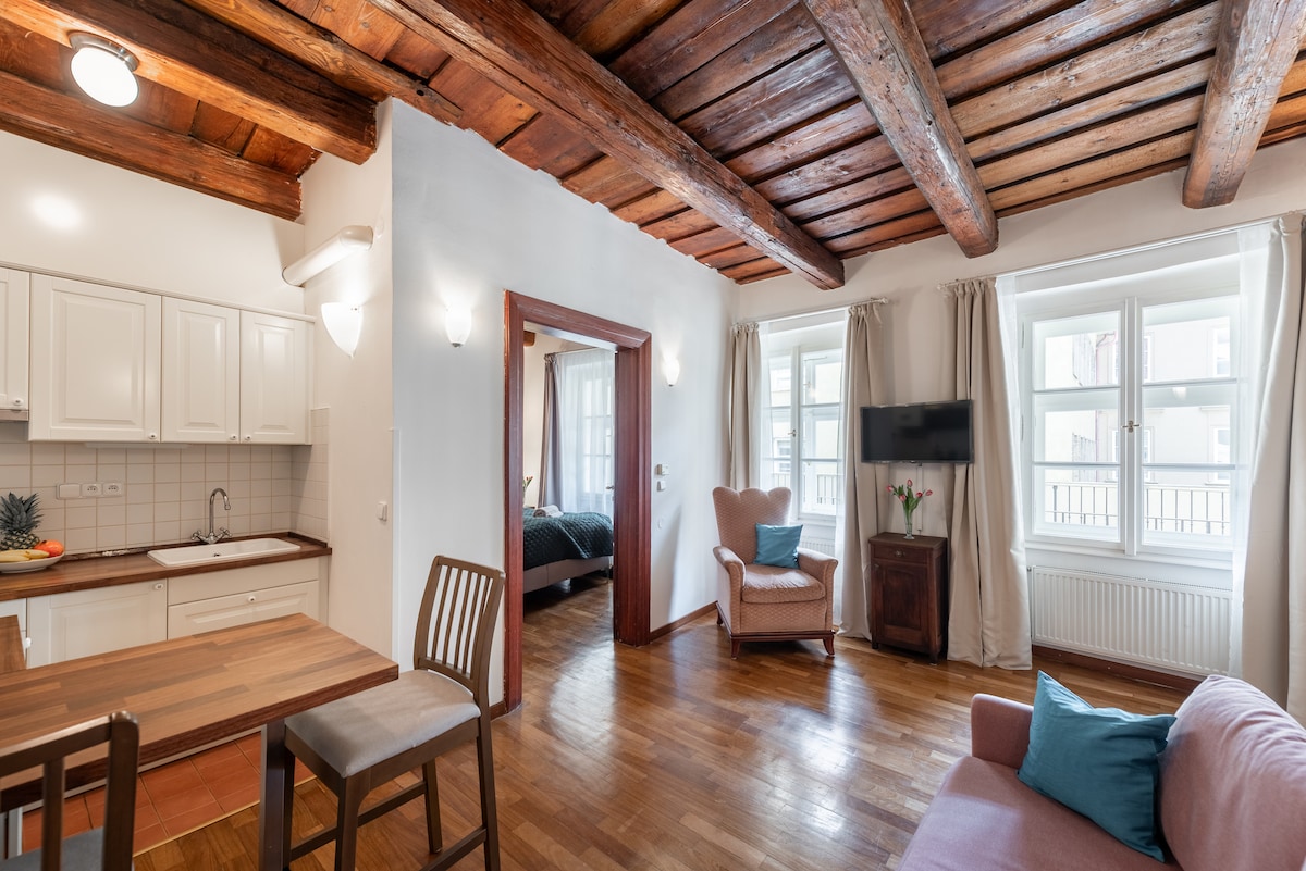 This spacious living area features wooden beams across the ceiling, complemented by large windows that allow natural light to fill the room. A dining table sits adjacent to a modern kitchen, while a cozy seating arrangement includes an armchair and a sofa, providing comfort and relaxation.