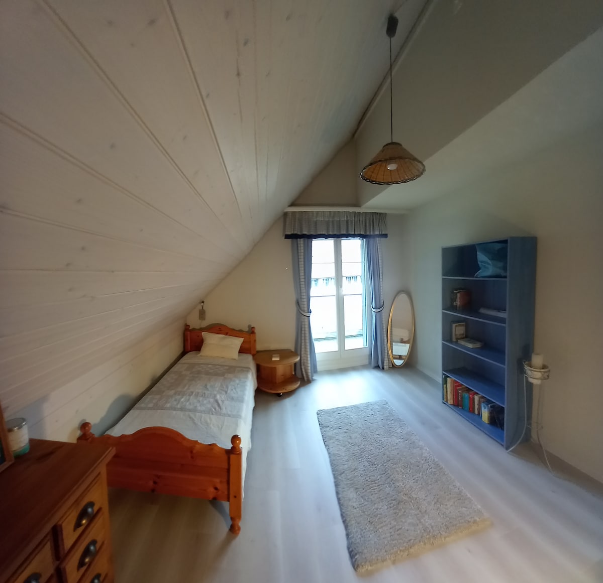 A cozy bedroom features a single wooden bed alongside a nightstand. A full-length mirror leans against the wall, and a bookshelf holds a few decorative items. Natural light fills the room through a balcony door, which is draped with light curtains.