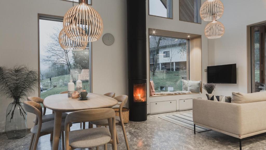 A modern living space features a round dining table with wooden chairs and decorative items. A cozy seating area is arranged around a stylish fireplace, with a large window nook showcasing outdoor views. Natural light floods the room through expansive windows, enhancing the inviting ambiance.