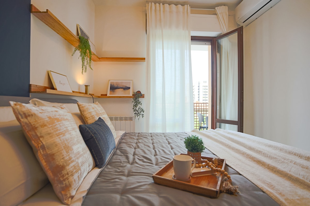 A softly lit bedroom features a spacious bed adorned with textured pillows. Light-colored curtains frame an open window, allowing natural light to brighten the space. Wall shelves hold decorative elements, while a small tray with a cup and greenery adds a touch of comfort.