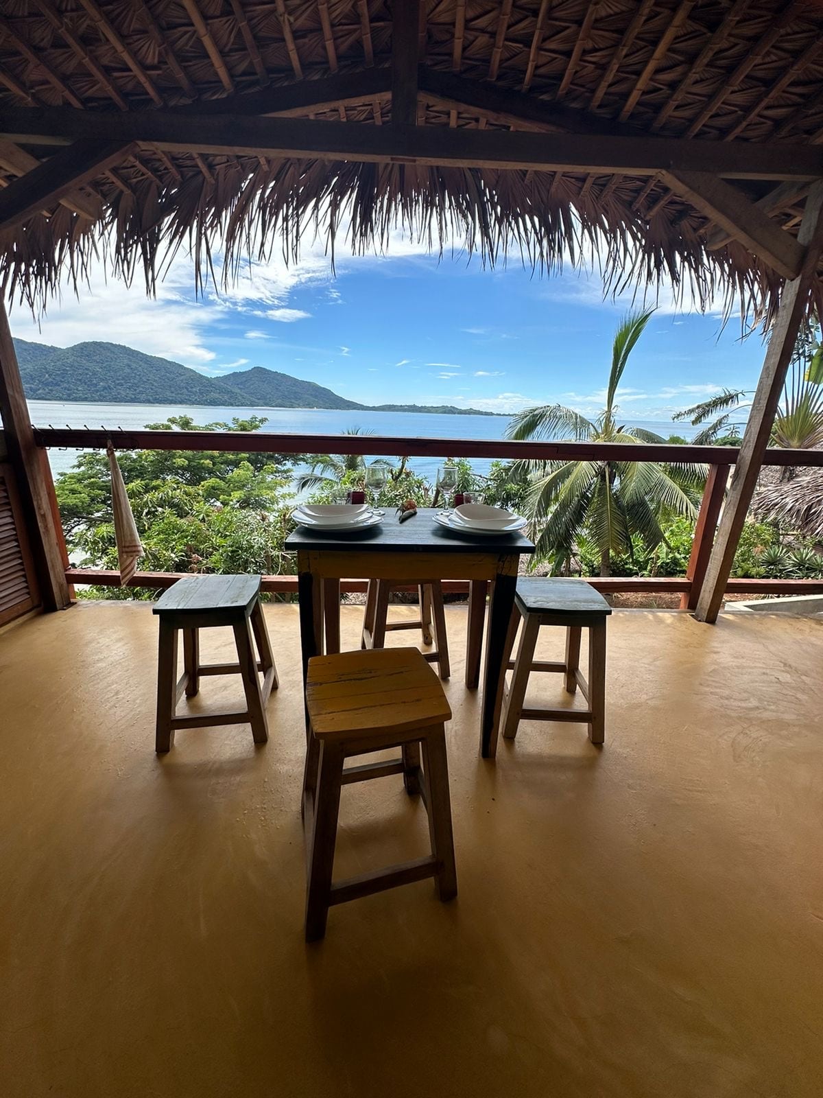 A dining area features a small table with two bowls and four wooden stools, all positioned on a terrace. Large windows provide a view of distant mountains and palm trees, with the shimmering water visible beyond.