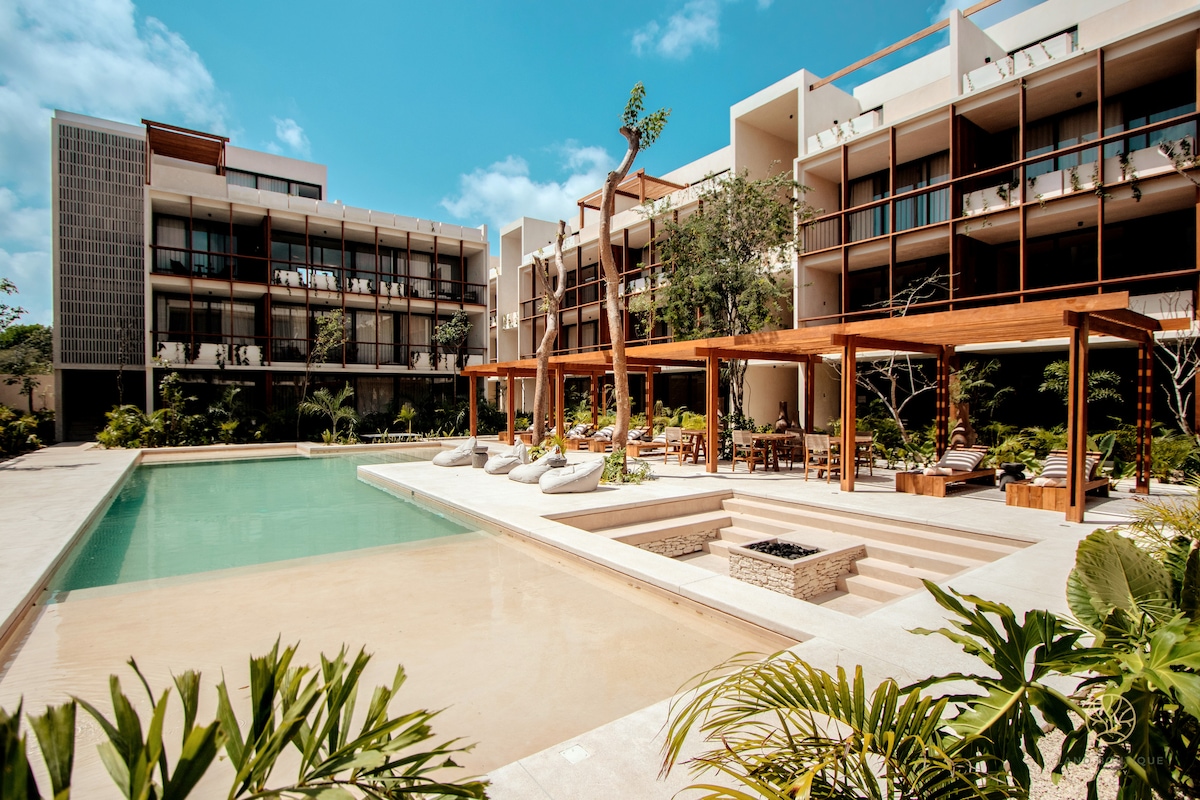 A private pool area is surrounded by landscaped greenery, with comfortable lounge seating under wooden pergolas. The modern building features large balconies and sliding glass doors, creating an open and inviting environment. Bright blue skies complement the serene atmosphere.
