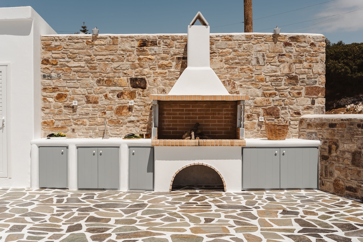An outdoor kitchen area features a stone wall with a built-in barbecue grill, complemented by a brick chimney. Below the grill, a countertop extends across the space, flanked by several storage cabinets in soft grey. The flooring consists of a patterned stone layout, enhancing the open atmosphere.