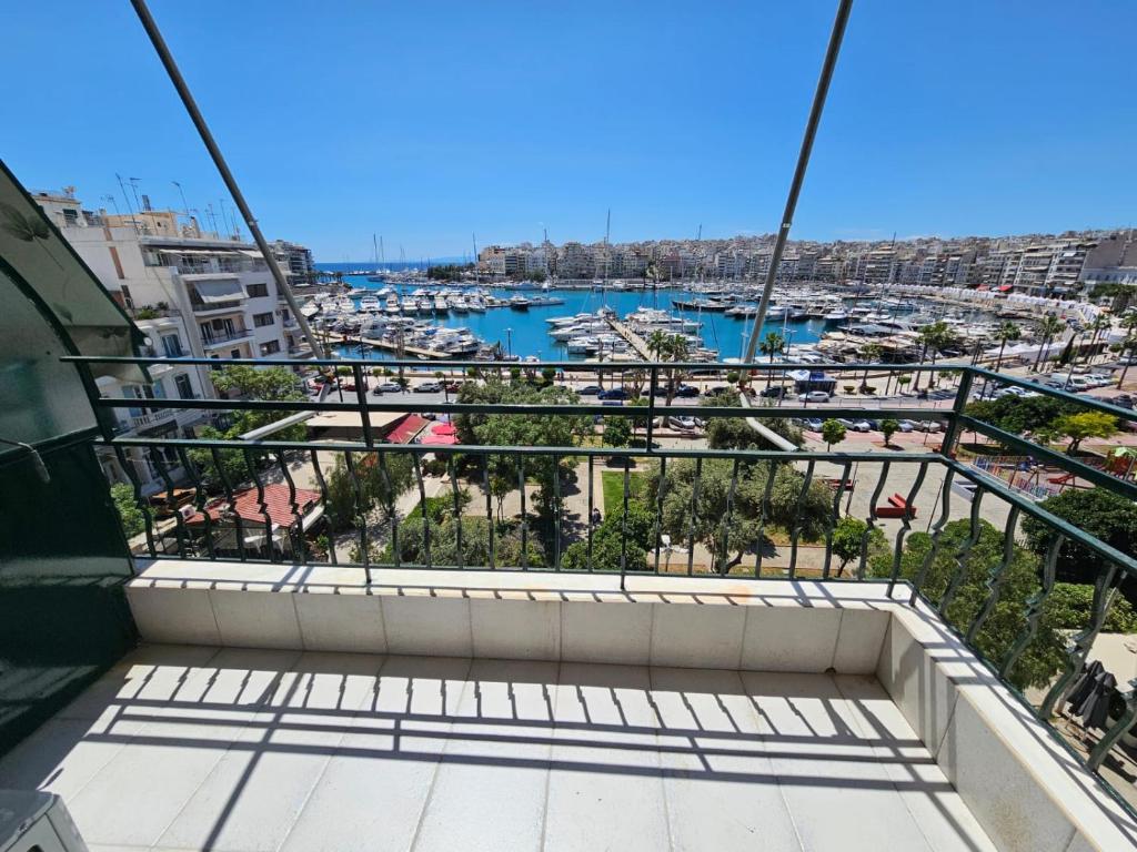 A spacious balcony offers a clear view of a bustling marina, filled with numerous boats and yachts. The vibrant blue sea contrasts with the surrounding greenery and urban landscape, while bright sunlight casts distinct shadows across the tiled floor.