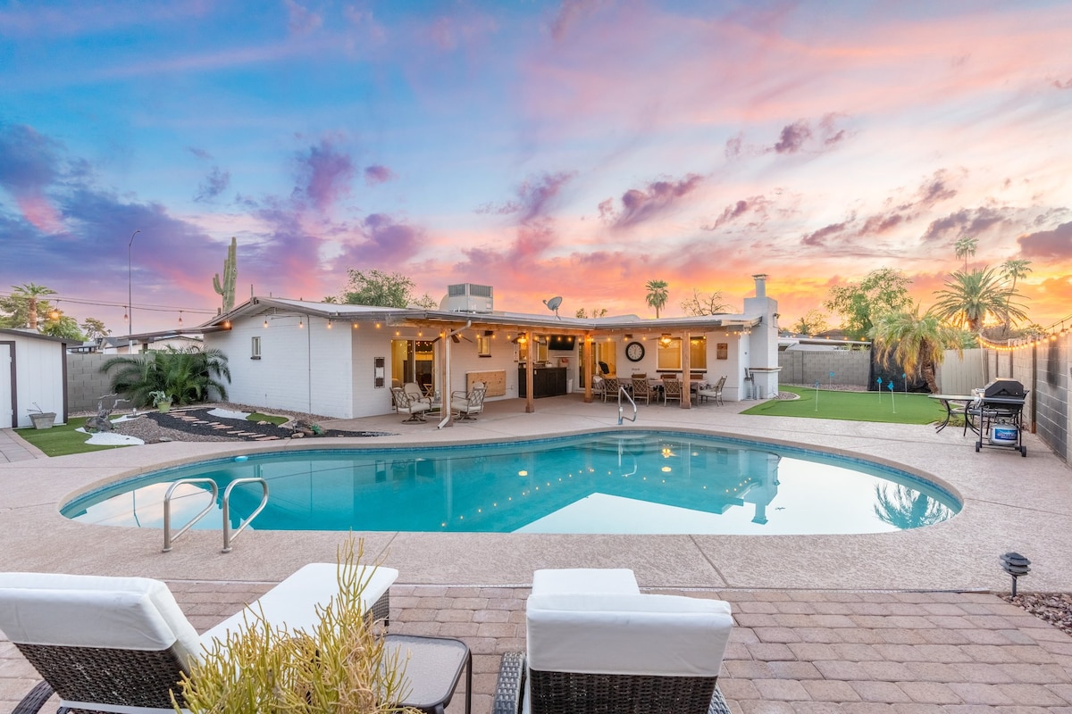 A serene pool area is viewed, featuring a refreshing pool surrounded by modern landscaping. Nearby, comfortable loungers are positioned for relaxation, while the home's exterior is visible against a colorful sunset sky, showcasing an inviting atmosphere for outdoor enjoyment.