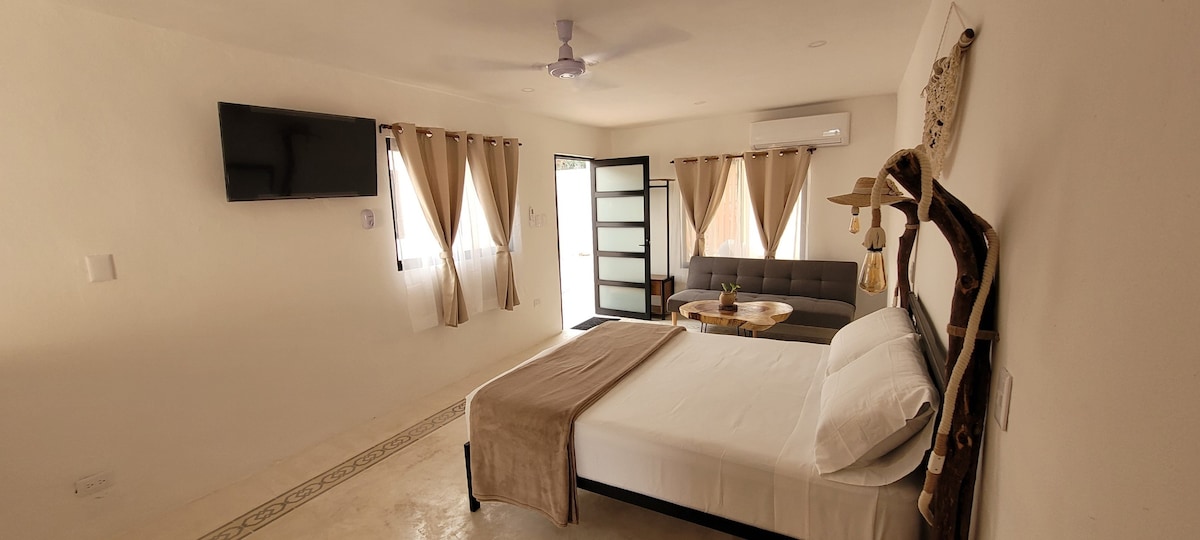 A well-appointed suite features a spacious bed and a comfortable sofa, accompanied by a small table. Natural light flows through the windows, complemented by soft curtains. A flat-screen TV is mounted on the wall, enhancing the modern and functional design of the room.