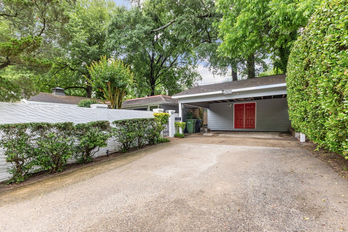 A private driveway leads to a carport, surrounded by lush greenery and low bushes. A white fence provides a sense of privacy. The carport features a distinctive red door, adding a pop of color to the serene outdoor environment.