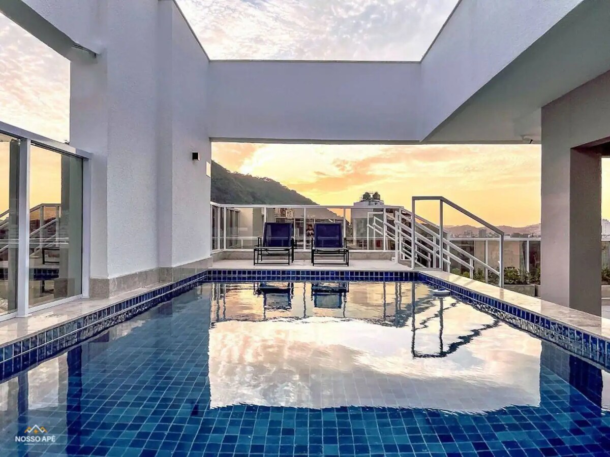A rooftop pool features a reflective blue surface, bordered by modern tiles. Two lounge chairs are positioned nearby, overlooking a scenic view framed by surrounding buildings. Soft sunlight casts a warm glow over the area, enhancing the tranquil setting.