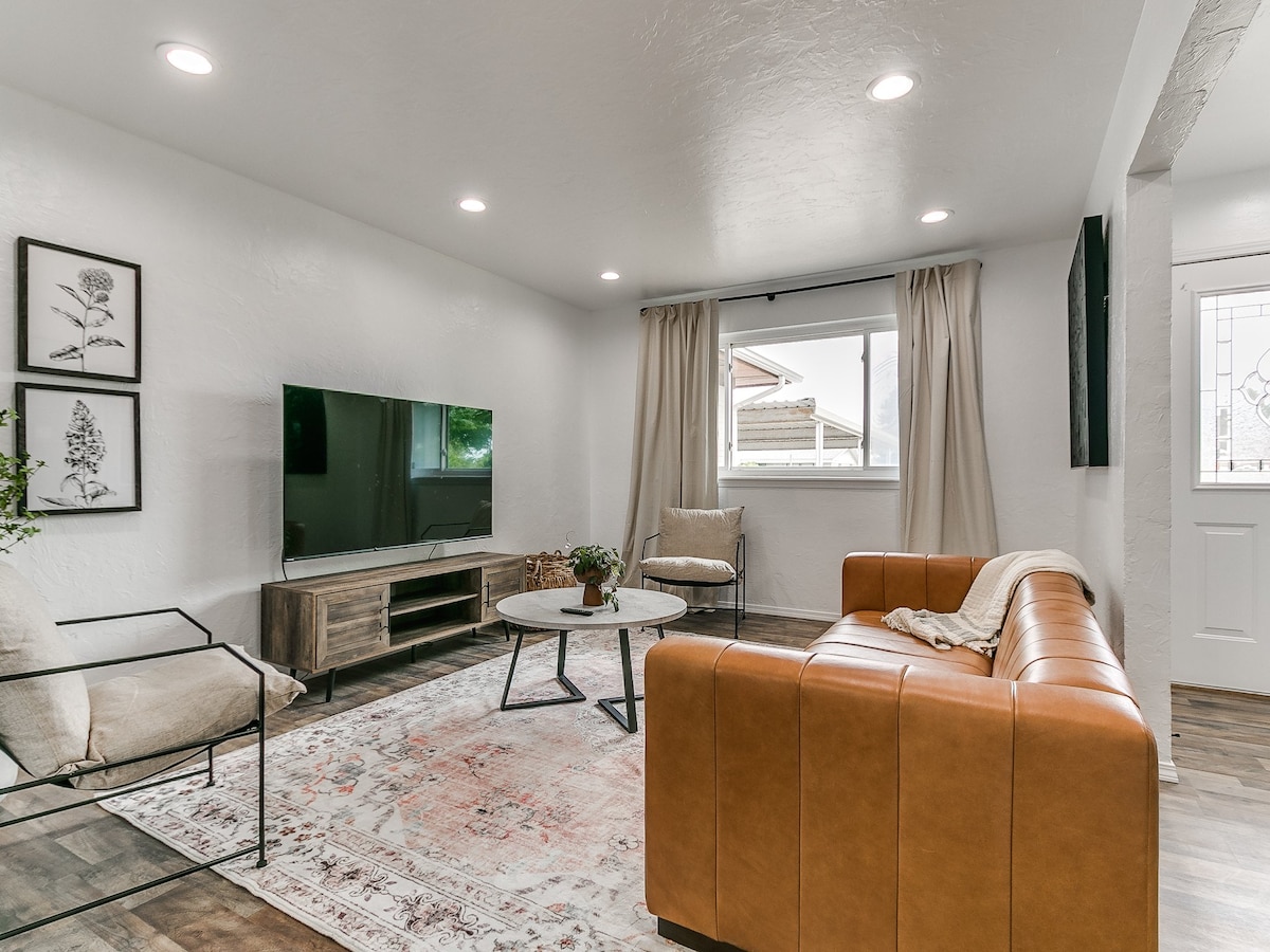 A modern living area features a large window allowing natural light to illuminate the space. A comfortable leather sofa is paired with a stylish coffee table on a patterned area rug. A wall-mounted television and decorative plants enhance the inviting layout.