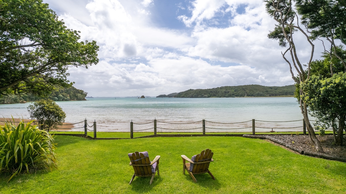 A serene view of the tidal beachfront is showcased, with two wooden chairs positioned on the lush green lawn. The inviting water reflects the clouds above, framed by distant hills and native trees along the shore.