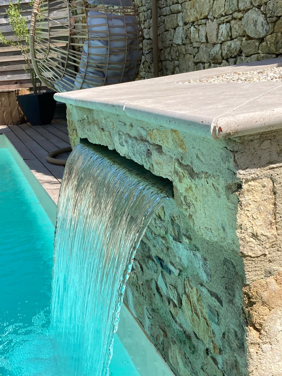 A stone water feature spills into a clear swimming pool, creating a serene ambiance. The vibrant turquoise water reflects the sunlight, enhancing the tranquil atmosphere. Stone walls in the background provide a natural backdrop, while potted greenery adds a touch of nature.