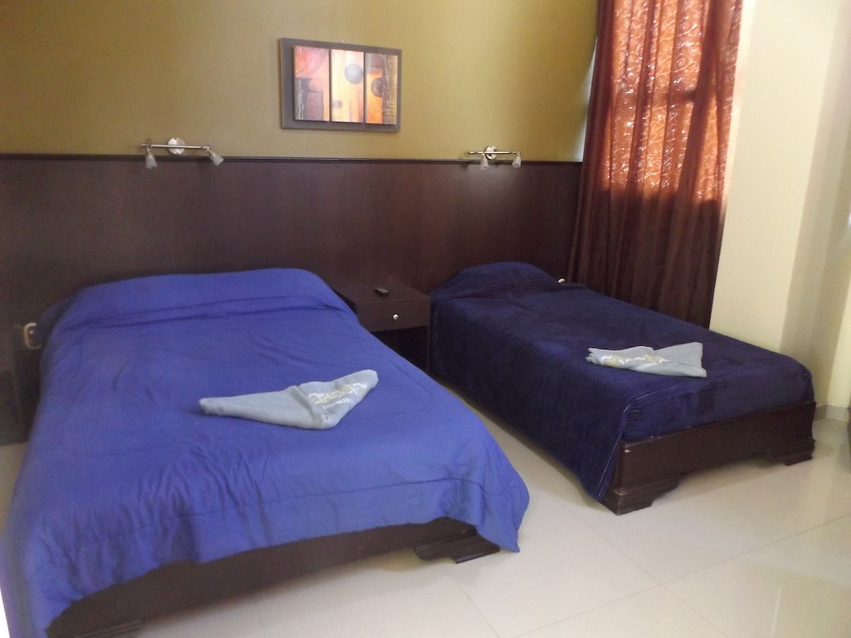 A well-lit room features two beds with blue linens, side tables, and wall-mounted reading lights. A window with light curtains allows natural light to enter, and a piece of artwork adds a decorative touch to the wall.