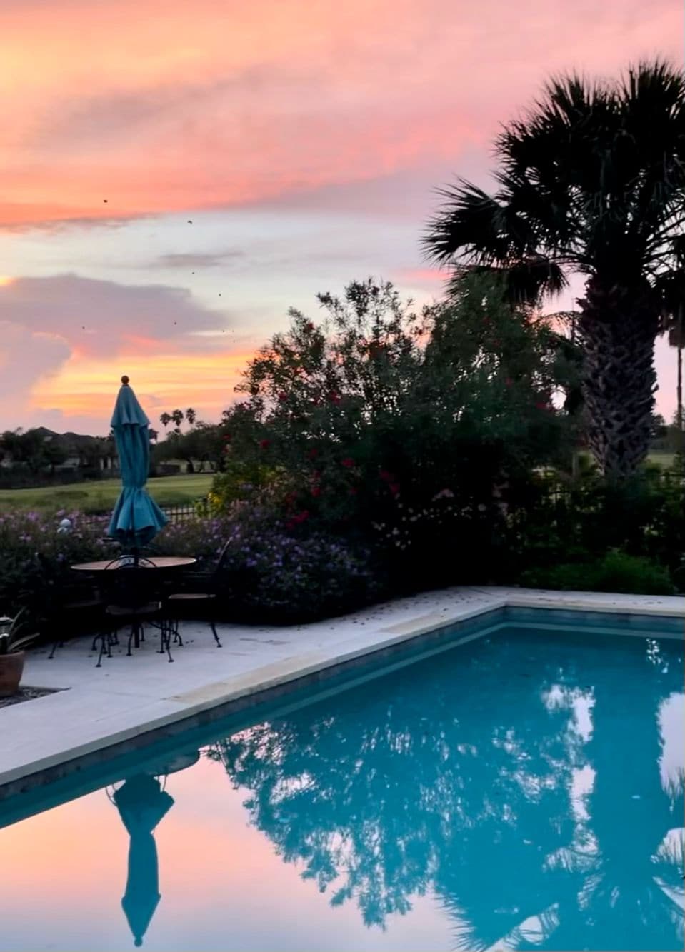 The serene outdoor setting features a private pool with crystal clear water, reflecting the vibrant hues of a sunset sky. Lush greenery and colorful flowers surround the pool area, complemented by a patio table and an umbrella, enhancing the tranquil ambiance.